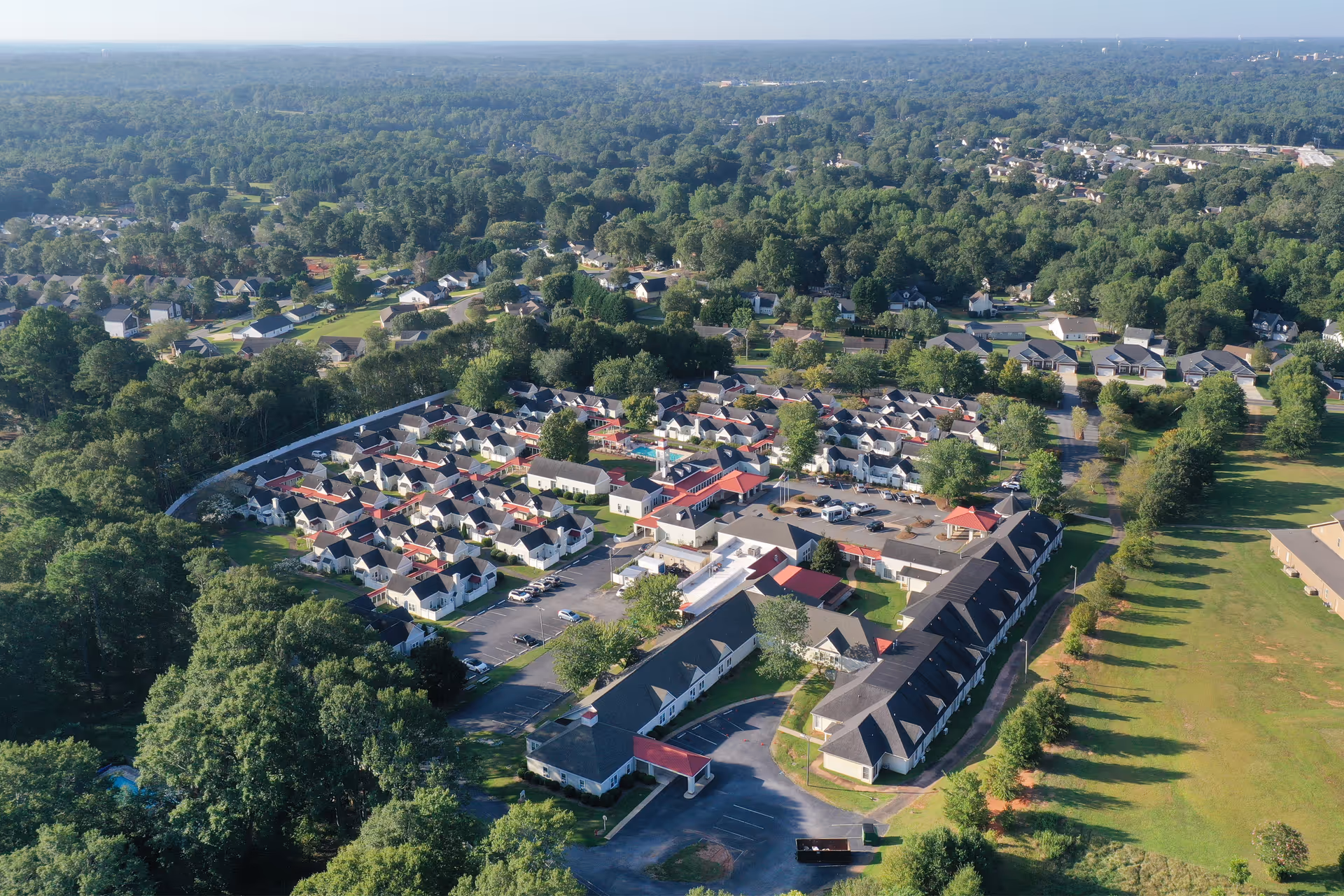 Aerial view of a senior living campus with rows of small residential cottages, central buildings, parking areas, and surrounding trees and lawns.