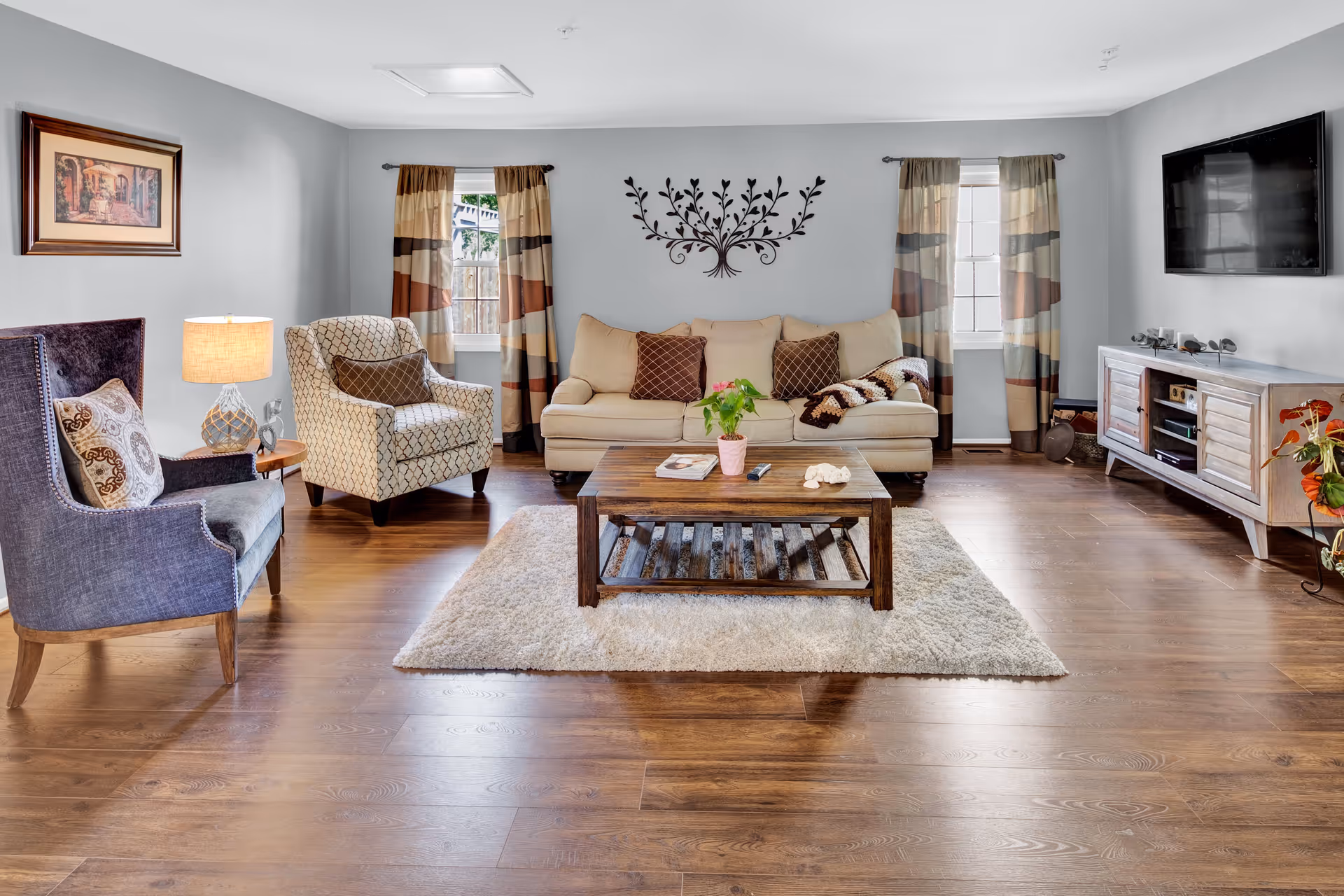 A cozy living room with a beige sofa adorned with brown pillows and a knitted throw, two patterned armchairs, a wooden coffee table with a plant and magazines on a light-colored rug, hardwood floors, two windows with striped curtains, a wall-mounted flat-screen TV, a wooden media console, and a decorative wall art piece above the sofa.