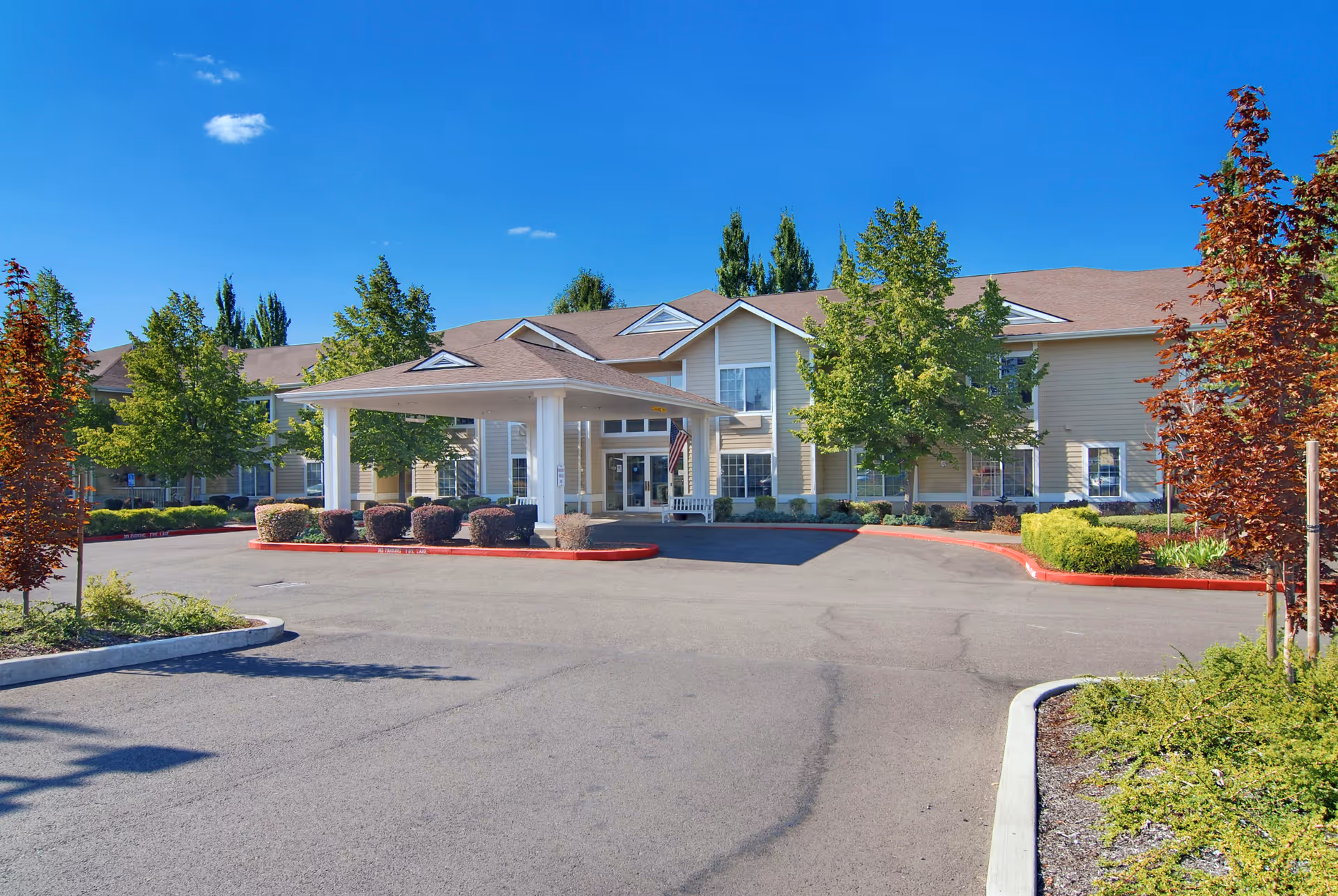 Front entrance of a two-story senior living building with a covered porte-cochere, landscaped grounds, and a driveway.