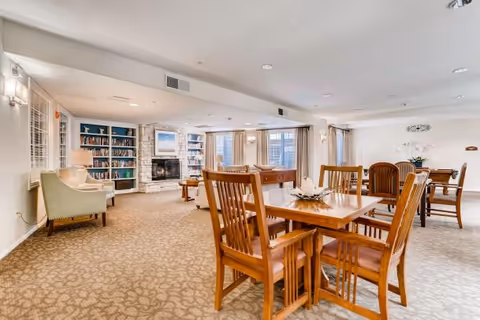 Bright communal lounge with wooden dining tables and chairs, a fireplace, bookshelves, and seating areas.