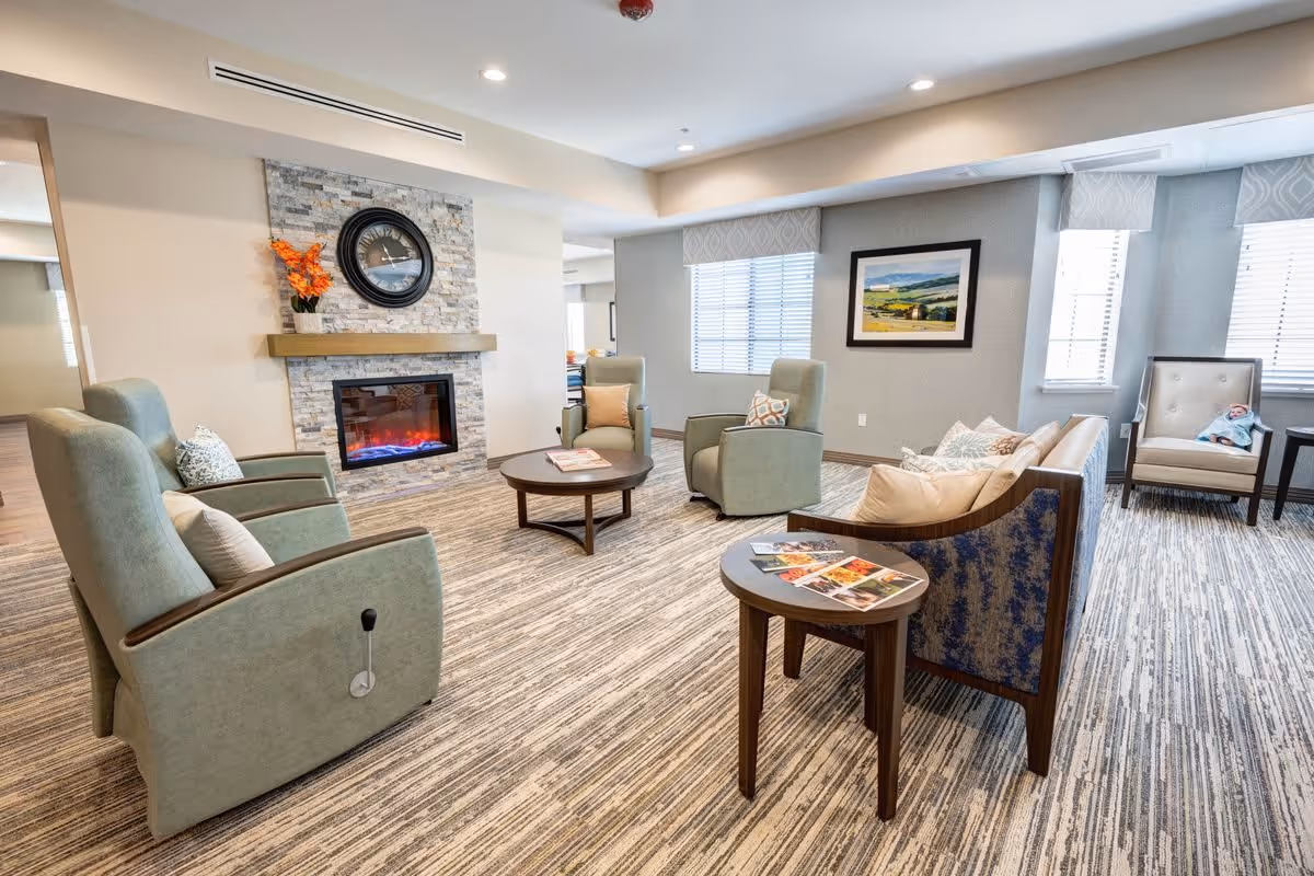 A cozy living room with a modern electric fireplace set in a stone wall with a clock above it. The room features several comfortable armchairs and a sofa arranged around two round wooden coffee tables. The space is well-lit with natural light from windows covered with blinds and light-colored walls decorated with a landscape painting. The carpet has a striped pattern, and there are some magazines on one of the tables.