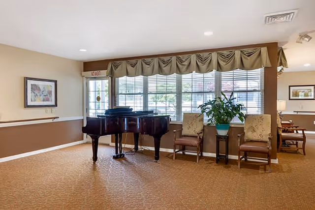 A spacious room with a grand piano in the center, two upholstered chairs on either side of a small table with a potted plant, large windows with blinds and valances letting in natural light, beige and brown walls, and carpeted floor.