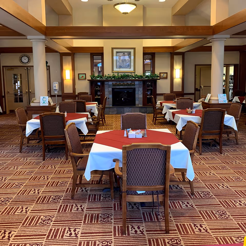 Spacious dining room with multiple tables covered in red and white tablecloths and wooden chairs arranged around a central fireplace.