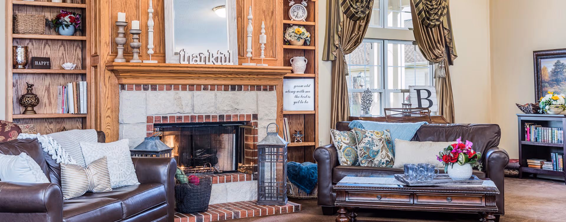 A cozy living room with two brown leather sofas adorned with decorative pillows, a wooden coffee table with a flower vase and glassware, a brick fireplace with candles and a 'thankful' sign on the mantel, wooden built-in shelves with books and decor, and large windows with brown curtains letting in natural light.