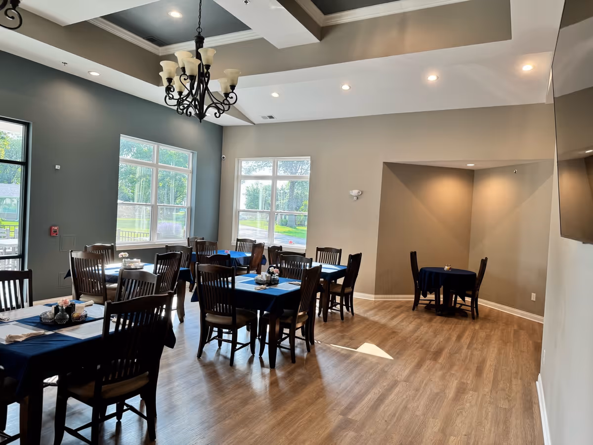 A bright dining room with multiple wooden tables and chairs arranged neatly. The tables are covered with dark blue tablecloths and have small decorative items on them. Large windows allow natural light to fill the room, and a decorative chandelier hangs from the ceiling. The floor is wooden, and the walls are painted in neutral tones.