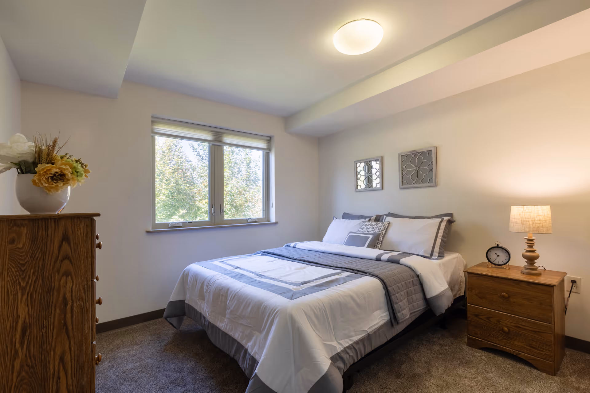 A neatly made bed with white and gray bedding in a bedroom with beige walls. There is a wooden nightstand with a lamp and a clock on it to the right of the bed, and a wooden dresser with a vase of flowers to the left. A window with a view of trees is centered on the wall behind the bed, and two decorative wall hangings are above the headboard.