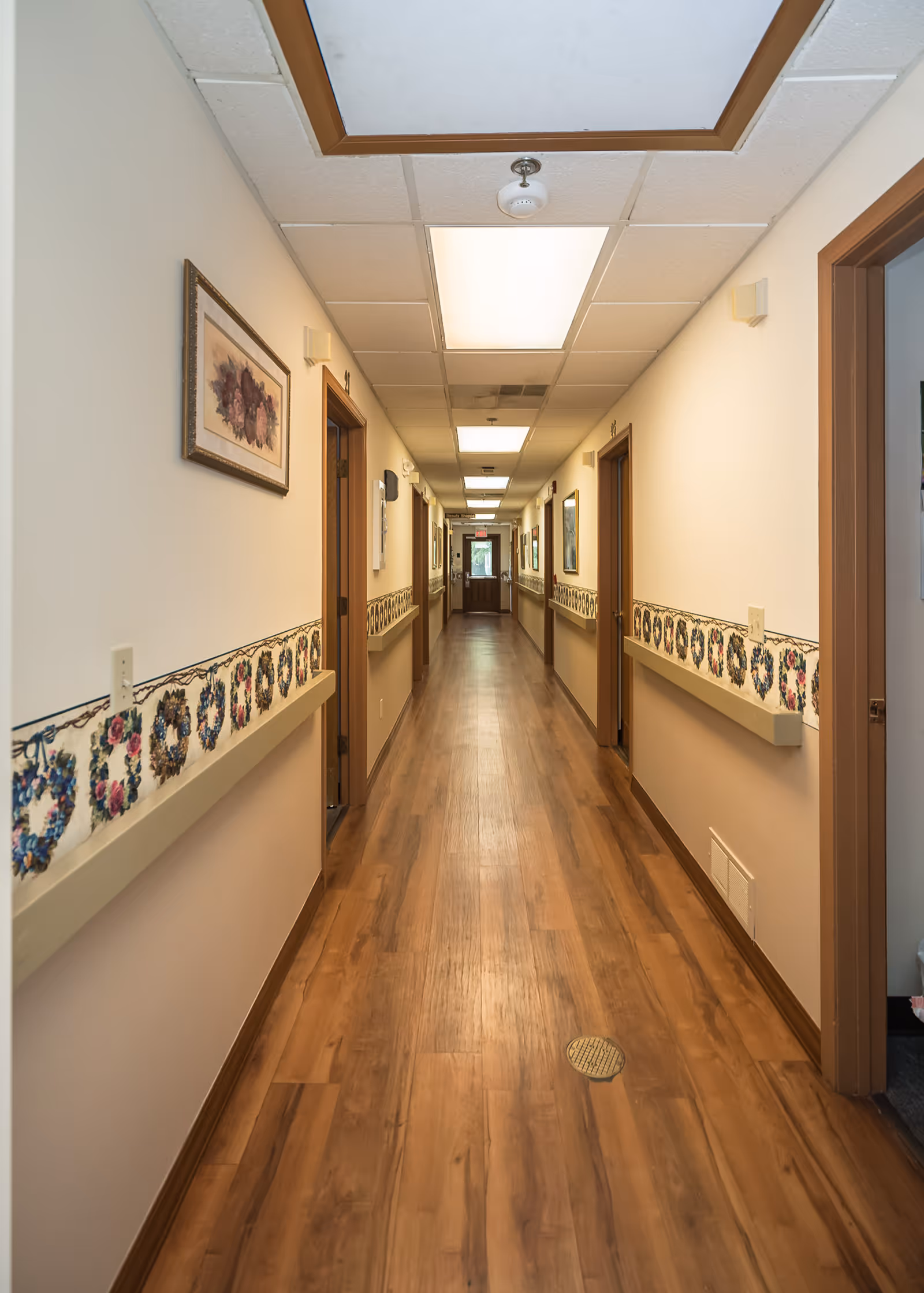 A long, well-lit hallway with wooden flooring and handrails on both sides decorated with floral patterns. Several doorways line the corridor, and framed pictures hang on the walls. The ceiling has recessed lighting panels and a smoke detector.