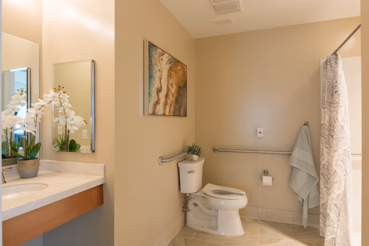 A clean and accessible bathroom with beige walls and tiled floor. The room features a white toilet with grab bars on the walls for support, a small potted plant on the toilet tank, a towel hanging on a hook, and a shower area with a patterned curtain. On the left side, there is a sink with a marble countertop, a mirror, and a decorative white orchid plant. A piece of abstract art hangs on the wall above the toilet.