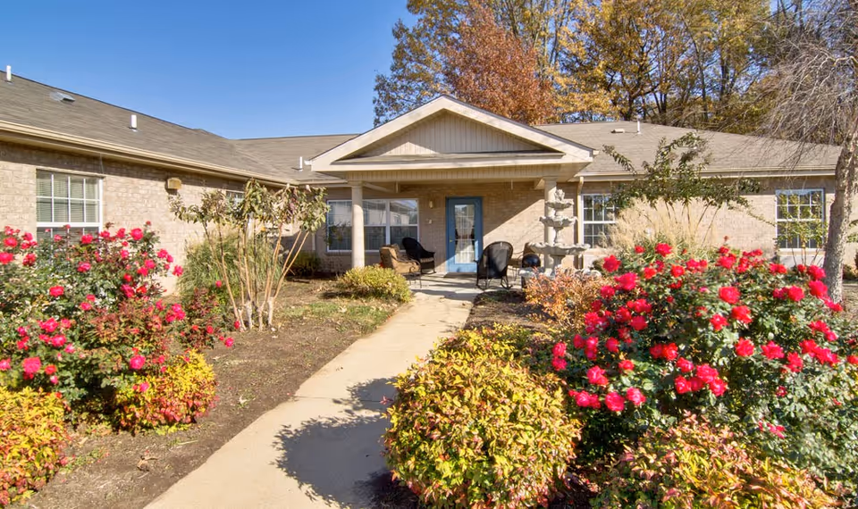 Exterior view of a single-story brick building with a covered entrance and a pathway leading to the door. The area is landscaped with bushes and vibrant red flowers under a clear blue sky.