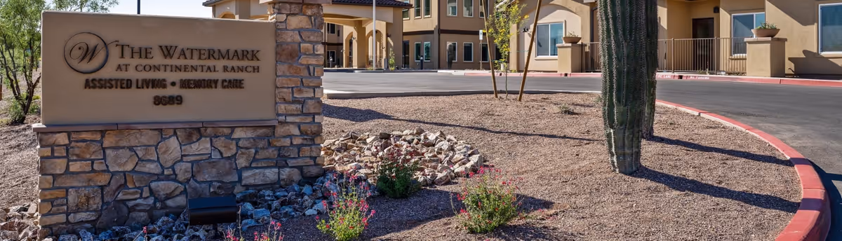 Entrance sign for The Watermark at Continental Ranch assisted living and memory care facility, with a stone base and beige sign. The background shows the facility building, desert landscaping with rocks, small plants, and a tall cactus under a clear sky.