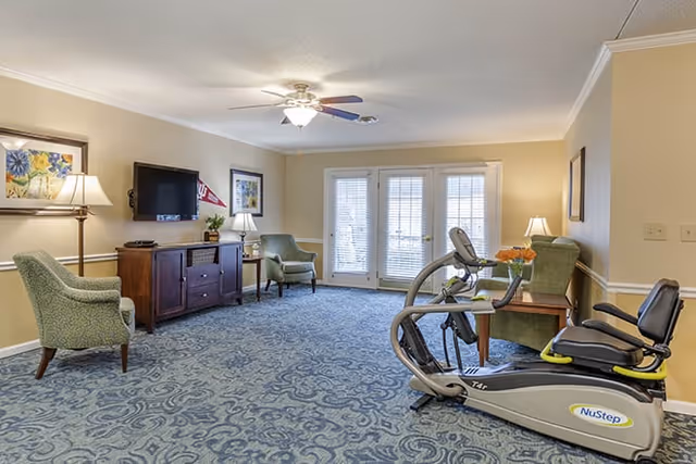 Bright common living room with armchairs, a wall-mounted TV, and a NuStep recumbent exercise machine near glass French doors.