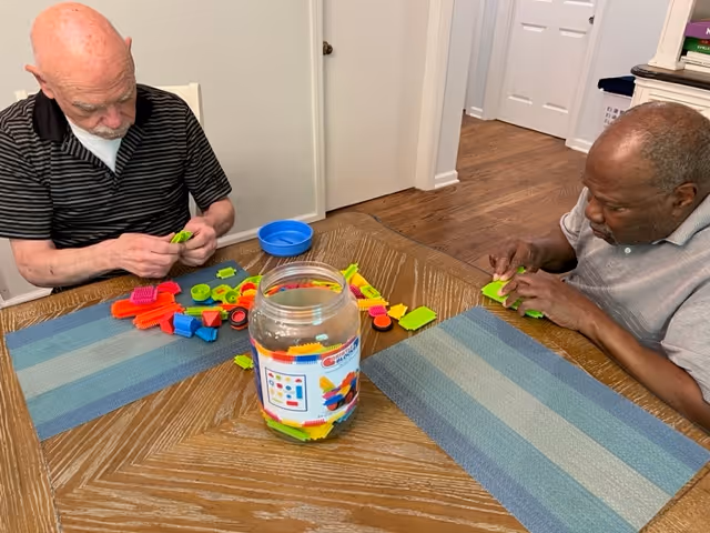 Two older men sit at a wooden table assembling colorful plastic construction pieces from a jar.