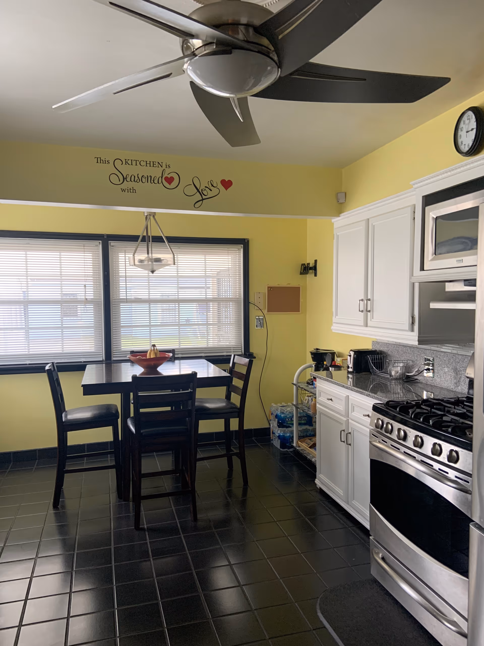 A kitchen with yellow walls, black tiled floor, and white cabinets. There is a stainless steel stove and microwave on the right side. A small dining table with four chairs is positioned near two large windows with blinds. Above the table, a hanging light fixture is visible. On the wall above the windows, there is a decorative sign that reads 'This KITCHEN is Seasoned with Love' with red heart accents. A ceiling fan with dark blades is mounted on the ceiling.