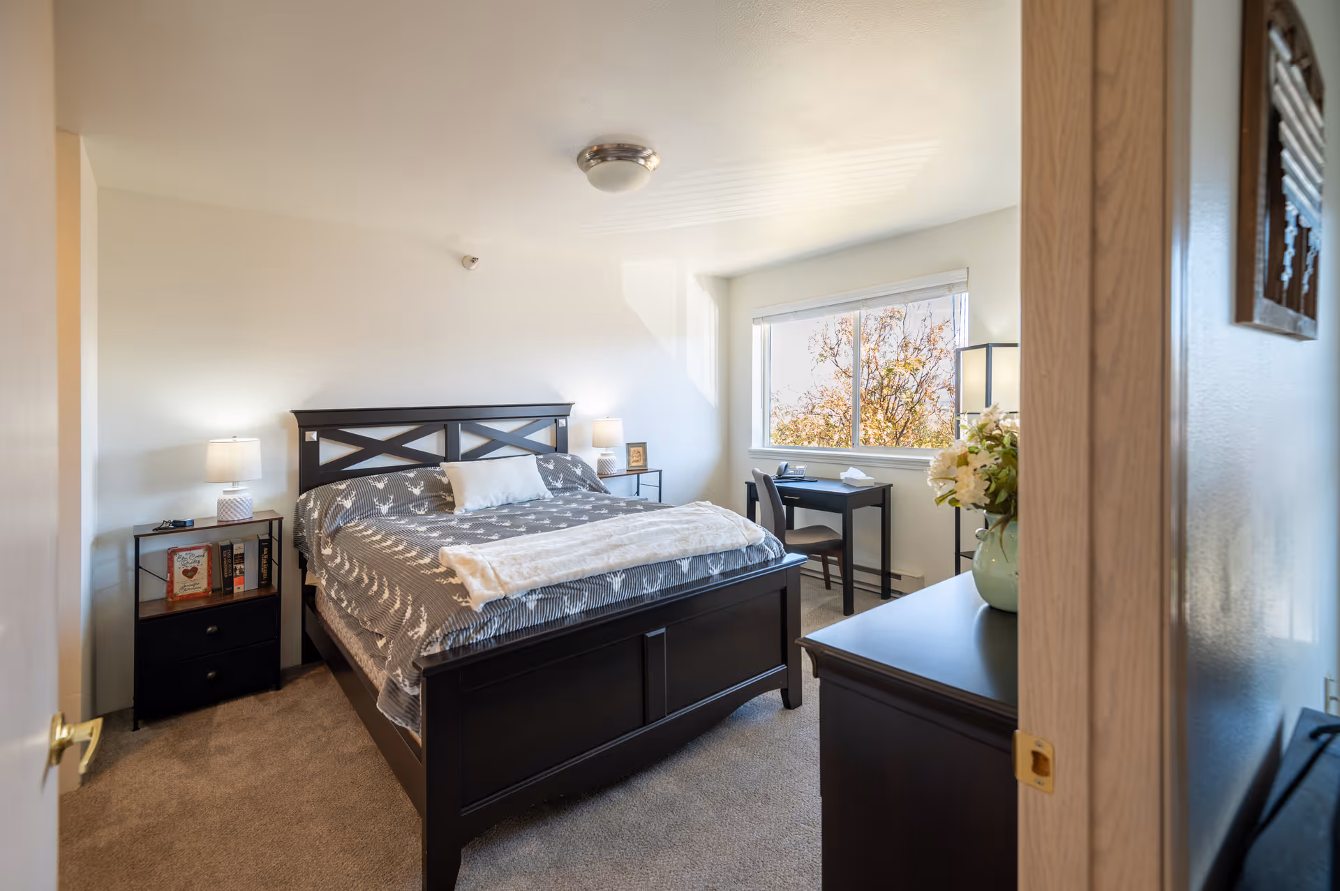 Well-lit furnished bedroom with a dark wood bed, nightstands, a desk by a window, and a dresser with flowers.