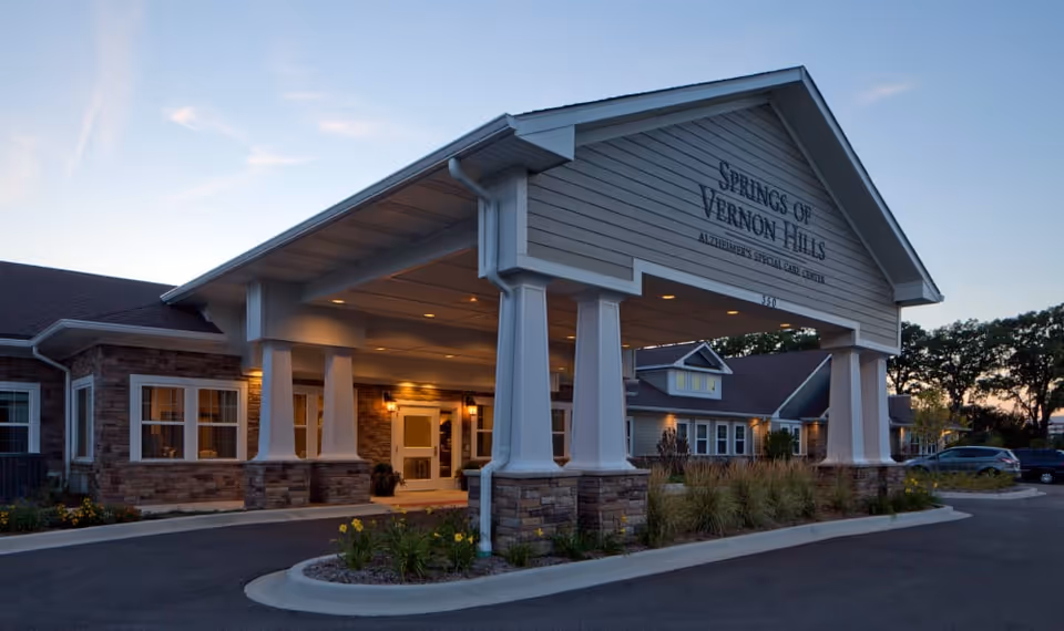Exterior view of the Springs of Vernon Hills Alzheimer's Special Care Center building at dusk, featuring a covered entrance with white columns and stone accents, surrounded by landscaping and a parking area.