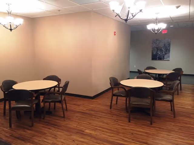 A room with wooden flooring featuring three round tables, each surrounded by four chairs. The walls are painted beige, and there are ceiling lights with multiple bulbs providing illumination. A framed picture hangs on the far wall near an exit sign.