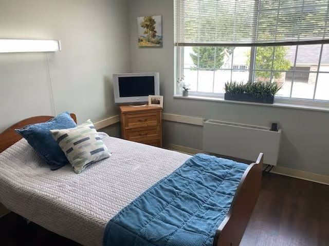 A small bedroom with a single wooden bed covered with a white quilt and blue throw blanket, two pillows, a wooden nightstand with a small TV and a framed photo, a large window with blinds, and a plant on the windowsill.