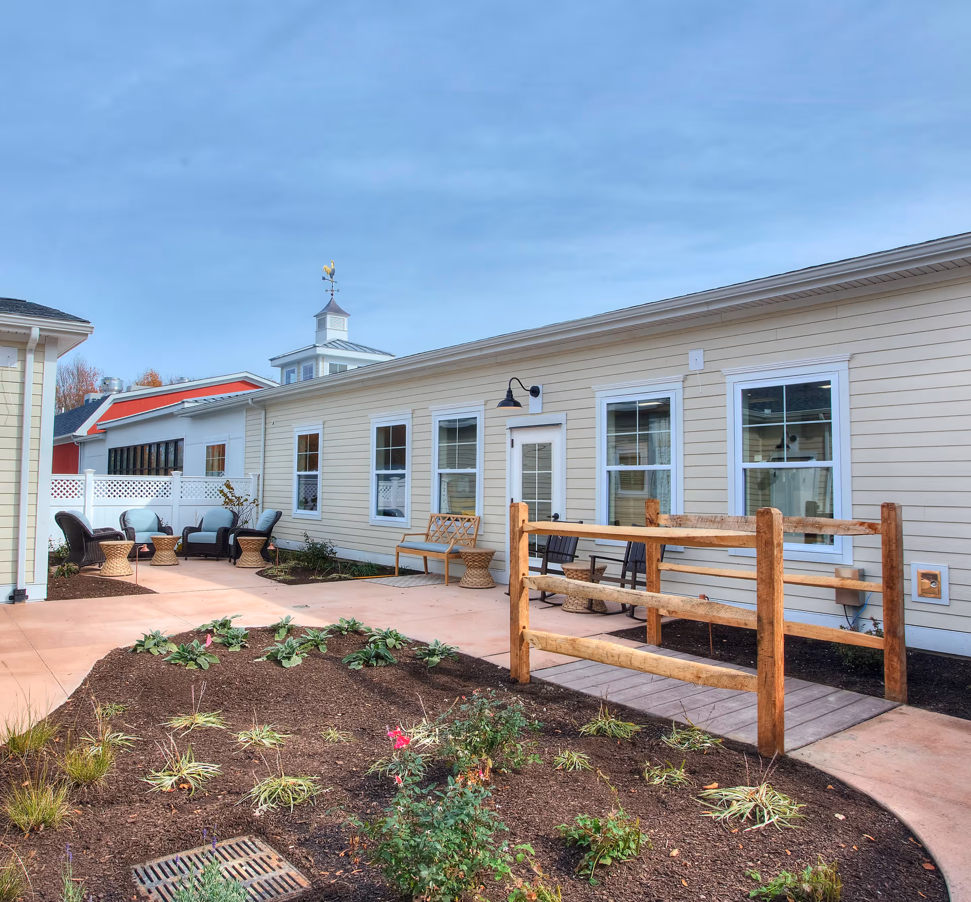 Outdoor patio area at Church Hill Village featuring a paved walkway, garden beds with plants, wooden railings, and several seating arrangements including cushioned chairs and small tables. The building exterior is light beige with multiple windows and a door, under a clear blue sky.