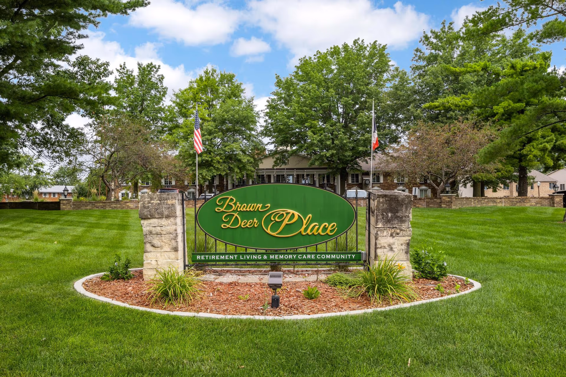 A green sign with gold lettering that reads 'Brown Deer Place' in front of a well-maintained lawn with trees and a building in the background. The sign also states 'Retirement Living & Memory Care Community'. Two flagpoles with flags are visible behind the sign.
