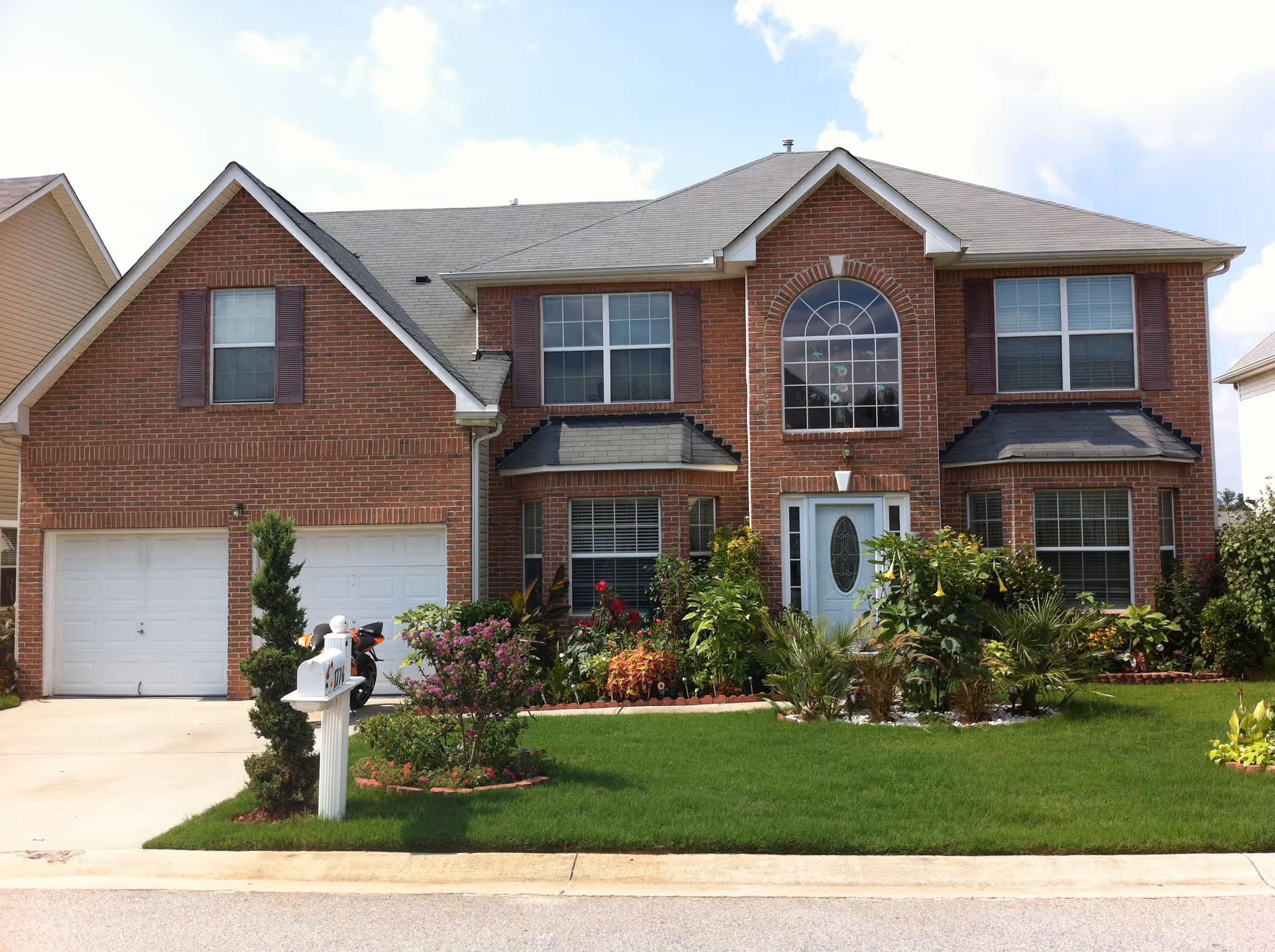 Front exterior view of a two-story brick house with a well-maintained lawn, various shrubs and flowering plants, a white mailbox, and a driveway leading to a double garage with white doors. The house has multiple windows with brown shutters and a white front door with an oval glass panel.