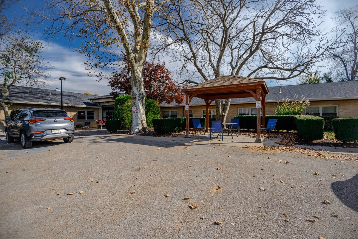 Outdoor area of Pennypack Nursing and Rehabilitation Center featuring a small wooden gazebo with blue chairs and a table underneath. There are trimmed bushes, leafless trees, and a parked gray SUV in the paved parking area. The building is a single-story structure with a brick exterior and several windows.