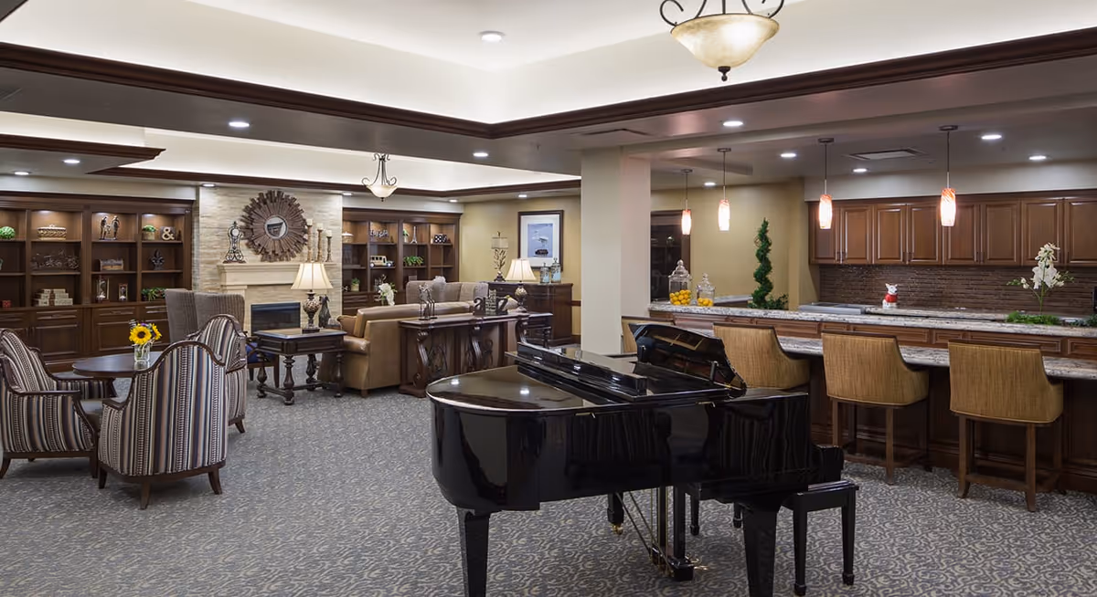 A spacious senior living common area featuring a black grand piano in the foreground, a kitchen bar with four wooden stools and pendant lighting, and a cozy seating area with armchairs, sofas, and built-in wooden bookshelves surrounding a fireplace.