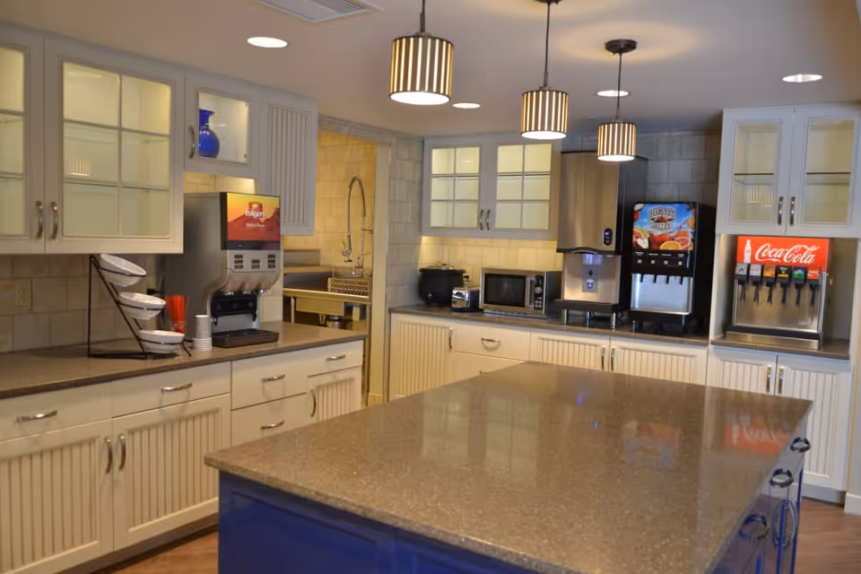 A modern kitchen area with white cabinets and a large central island with a polished countertop. The kitchen features multiple beverage dispensers including Folgers coffee, a juice machine, and a Coca-Cola soda fountain. There are pendant lights hanging from the ceiling and a microwave and toaster on the counter.