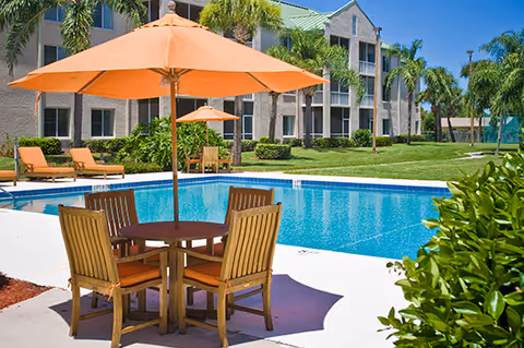 Outdoor swimming pool area with a round wooden table and four chairs under an orange umbrella. Several lounge chairs with orange cushions are placed near the pool. In the background, there is a multi-story residential building surrounded by palm trees and green grass.