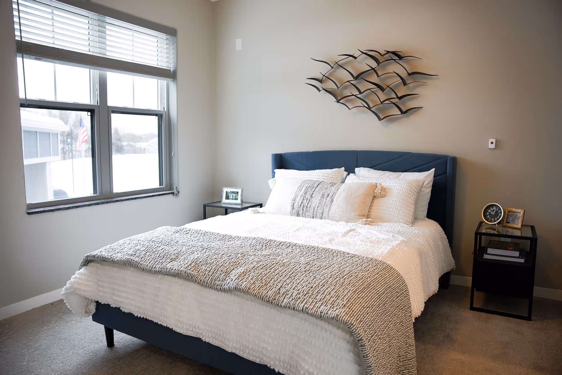 A neatly made bed with white and gray bedding in a bright bedroom. There is a large window with blinds on the left side, a black metal nightstand with a clock and picture frame on the right, and a decorative metal wall art above the bed.