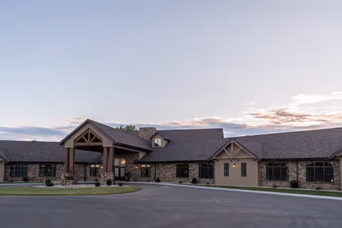 Exterior view of a single-story senior living facility building with a covered entrance, stone and siding facade, and a paved driveway under a partly cloudy sky at dusk.