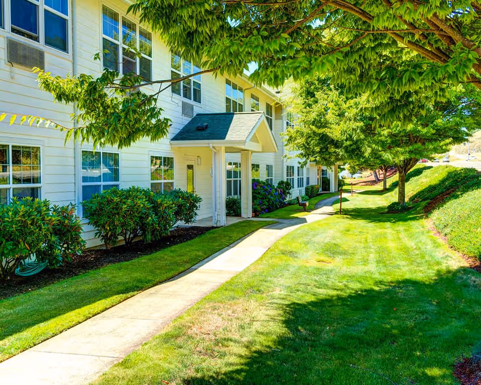 Exterior view of Callahan Village Retirement and Assisted Living building with a white facade, multiple windows, a small covered entrance, and a well-maintained lawn with trees and shrubs along a paved walkway.