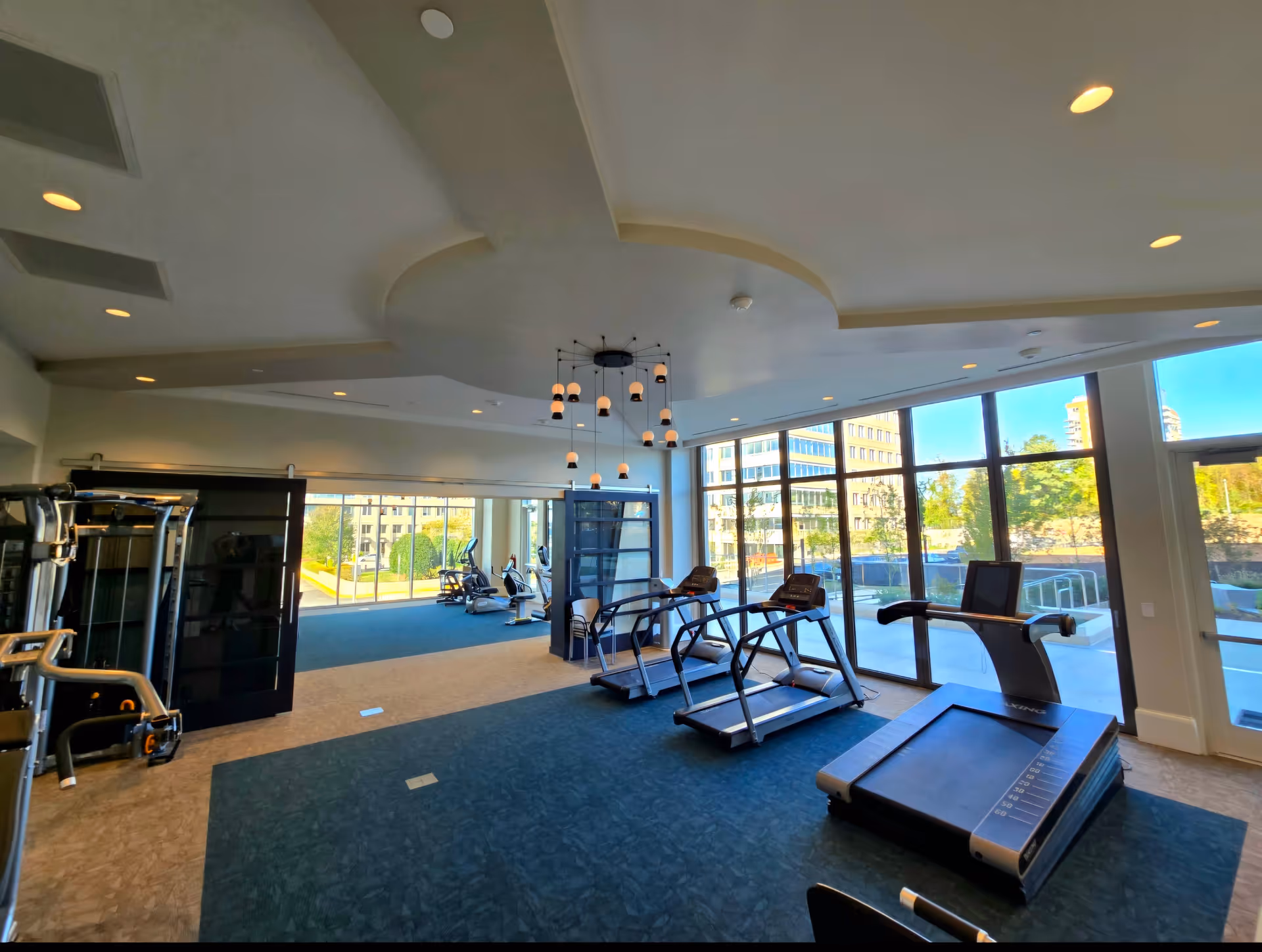 A bright fitness room with large windows showing an outdoor view of buildings and trees. The room contains several treadmills, exercise bikes, and other gym equipment. The ceiling features a modern circular design with hanging pendant lights.