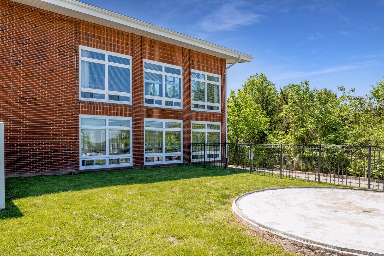 Exterior view of a brick building with large windows and a grassy area in front. There is a circular concrete pad on the grass and a black metal fence along the edge of the property. Trees and greenery are visible in the background under a clear blue sky.