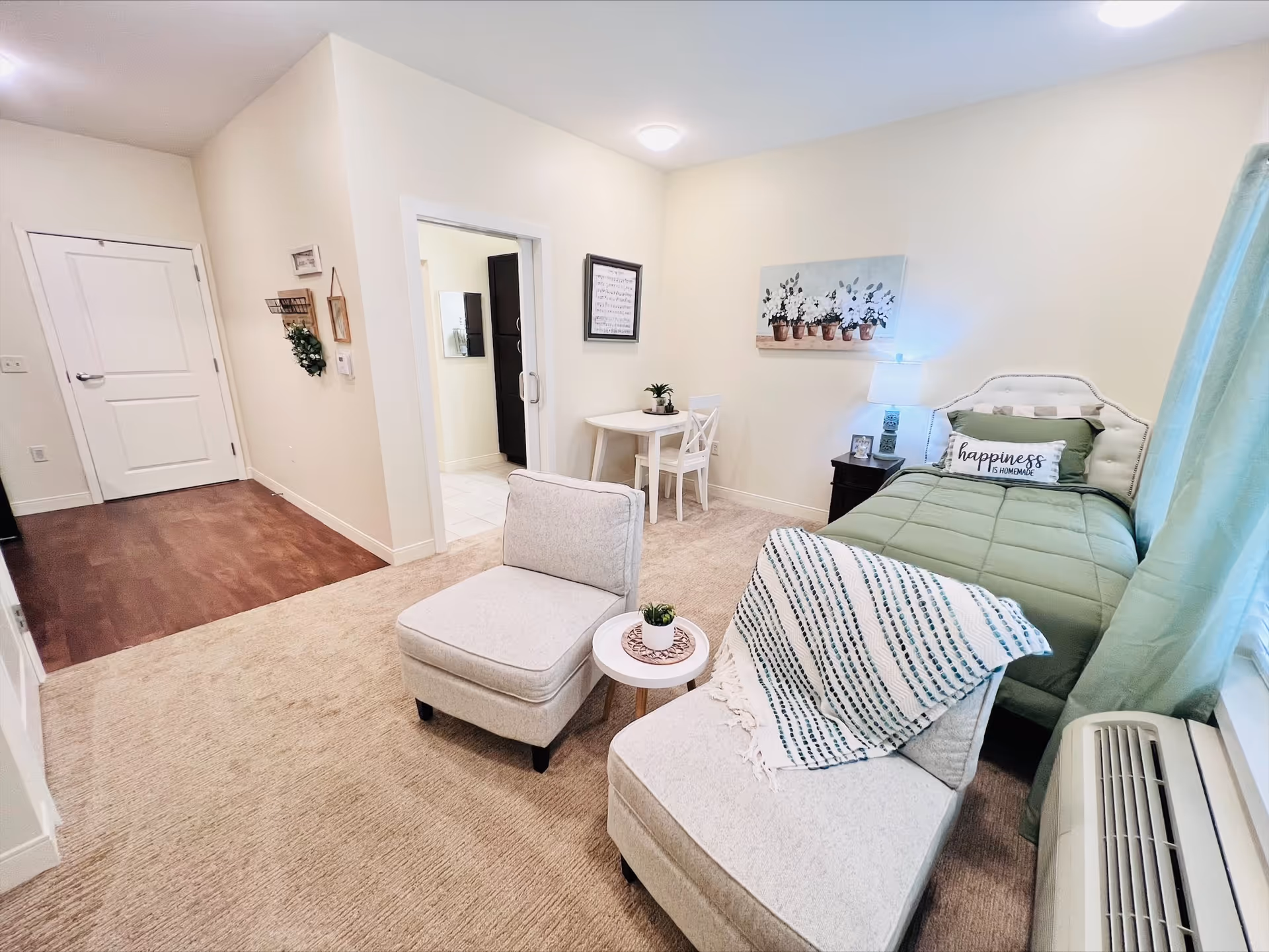 A cozy bedroom in an assisted living facility featuring a single bed with green bedding and a pillow that says 'happiness is homemade'. There are two cushioned chairs with a small round table between them, a small dining table with two chairs, a nightstand with a lamp, and wall art depicting potted plants. The room has beige carpet and light-colored walls, with a window covered by green curtains and an air conditioning unit below it. A doorway leads to another room with tile flooring.