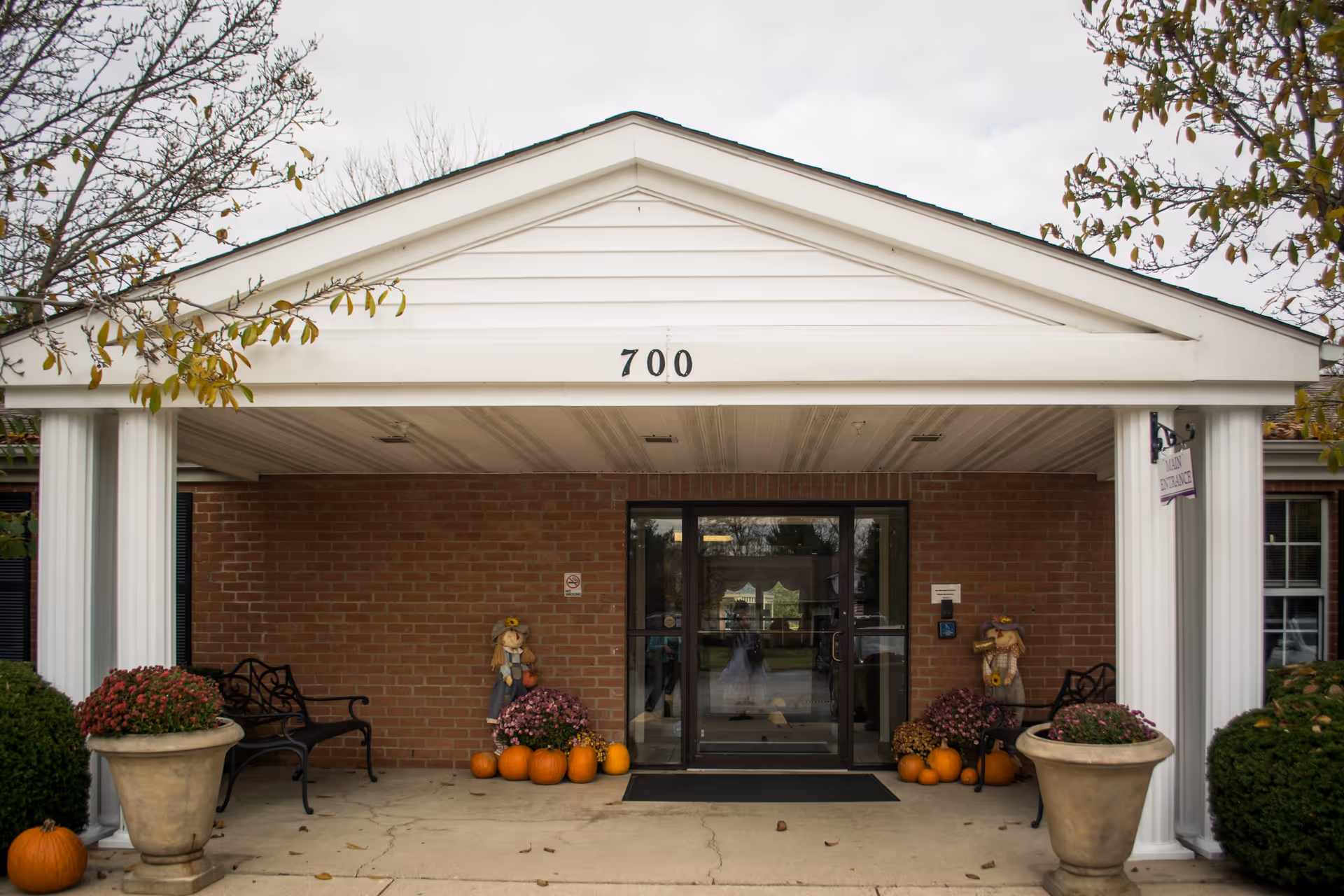 Front entrance of a brick building with the number 700 displayed above the doorway. The entrance is covered by a white portico with two white columns. There are two black benches on either side of the door, large flower pots with plants, and several pumpkins and autumn decorations near the entrance. Trees with some remaining leaves frame the scene.