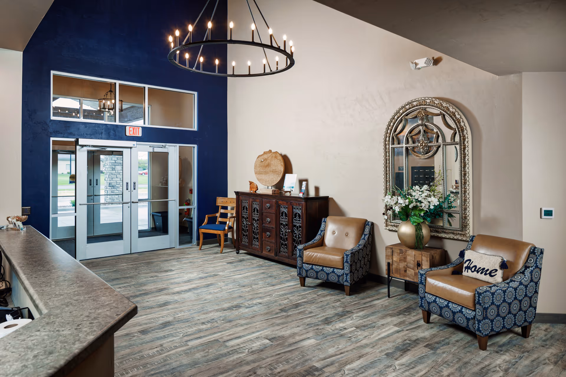 Lobby of a senior living facility showing double glass entrance doors, decorative chairs, a sideboard with mirror, and a chandelier.