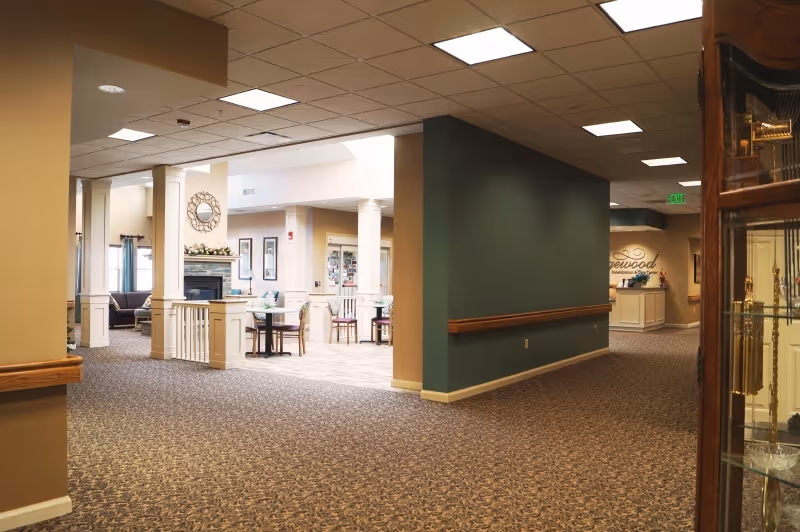 Well-lit, carpeted senior living facility interior showing a seating area with a fireplace, tables and chairs, columns, and a hallway.