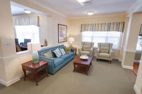 A cozy living room area with a blue sofa adorned with decorative pillows, two beige armchairs, a wooden coffee table with a white bowl, and a wooden side table with a lamp. The room has large windows with striped valances allowing natural light to fill the space. The walls are painted in a soft cream color with white trim.