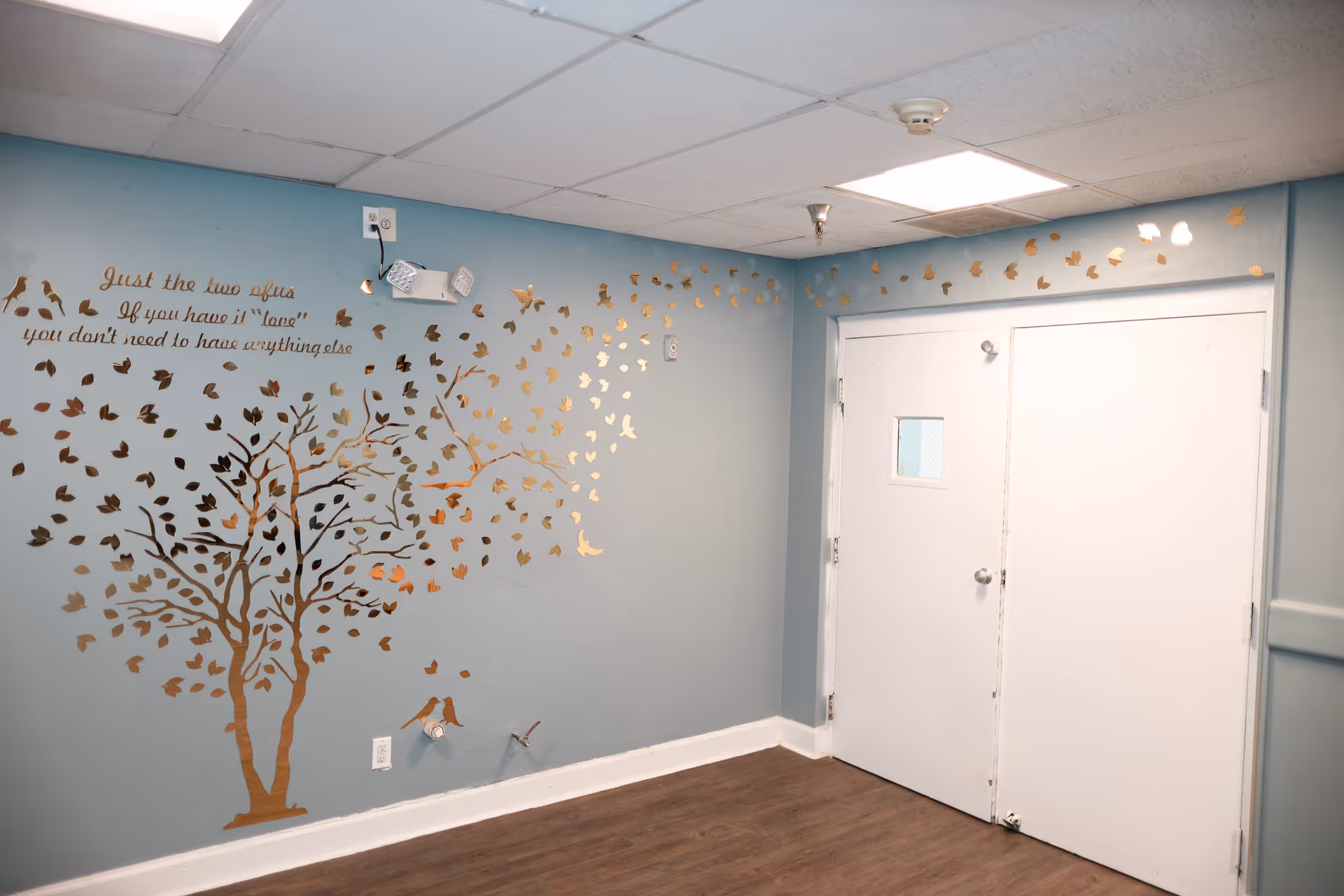Interior corner of a room with light blue walls and a white double door. The wall features a decorative gold tree decal with leaves and birds, along with a quote that reads, 'Just the two of us If you have it "love" you don't need to have anything else.' The floor is wood, and the ceiling has white tiles with fluorescent lighting.