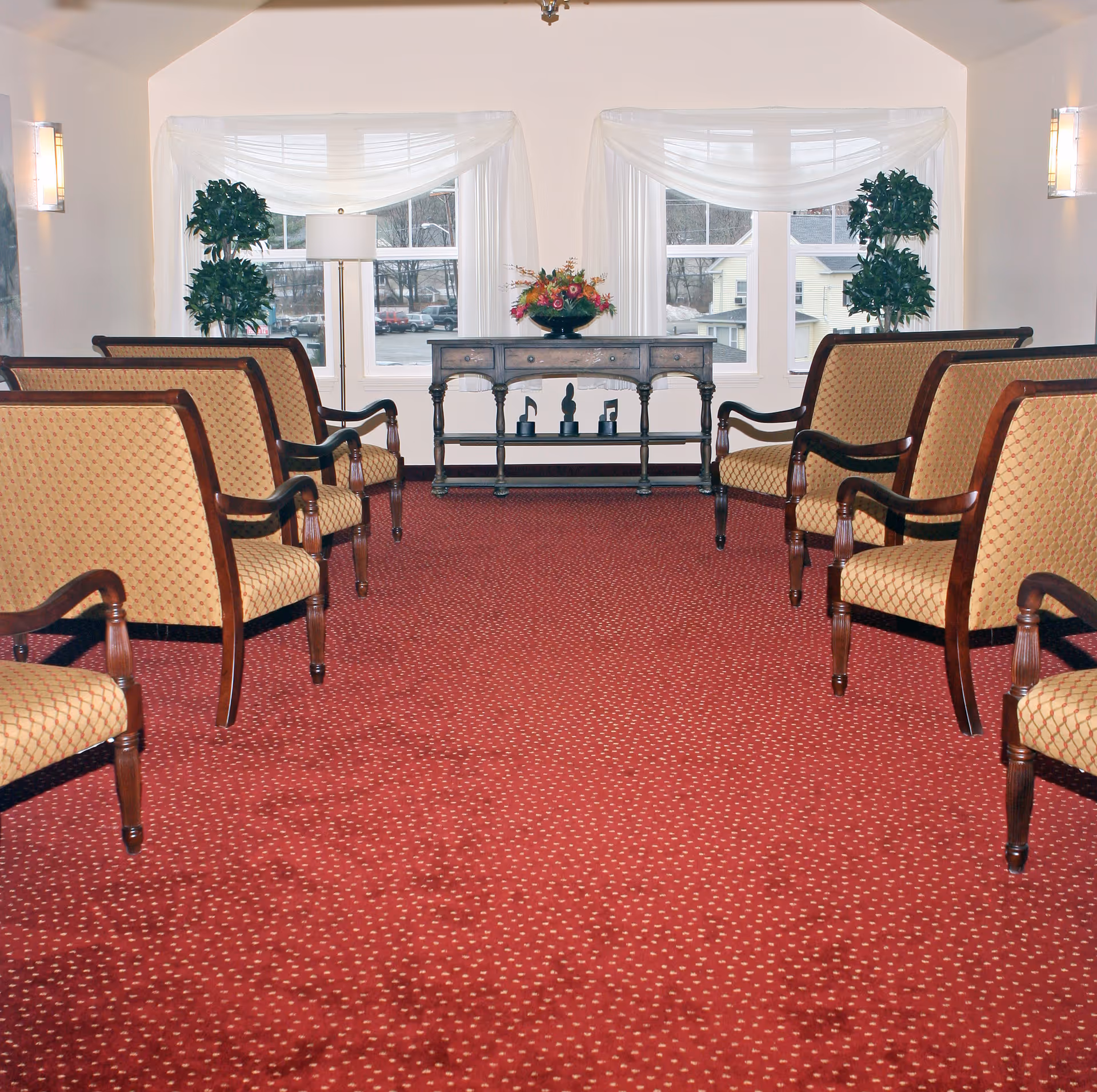 A sitting area with two rows of upholstered wooden chairs facing each other on a red patterned carpet. At the far end, there is a wooden console table with a flower arrangement and decorative items, flanked by two potted plants. Large windows with sheer white curtains allow natural light into the room.