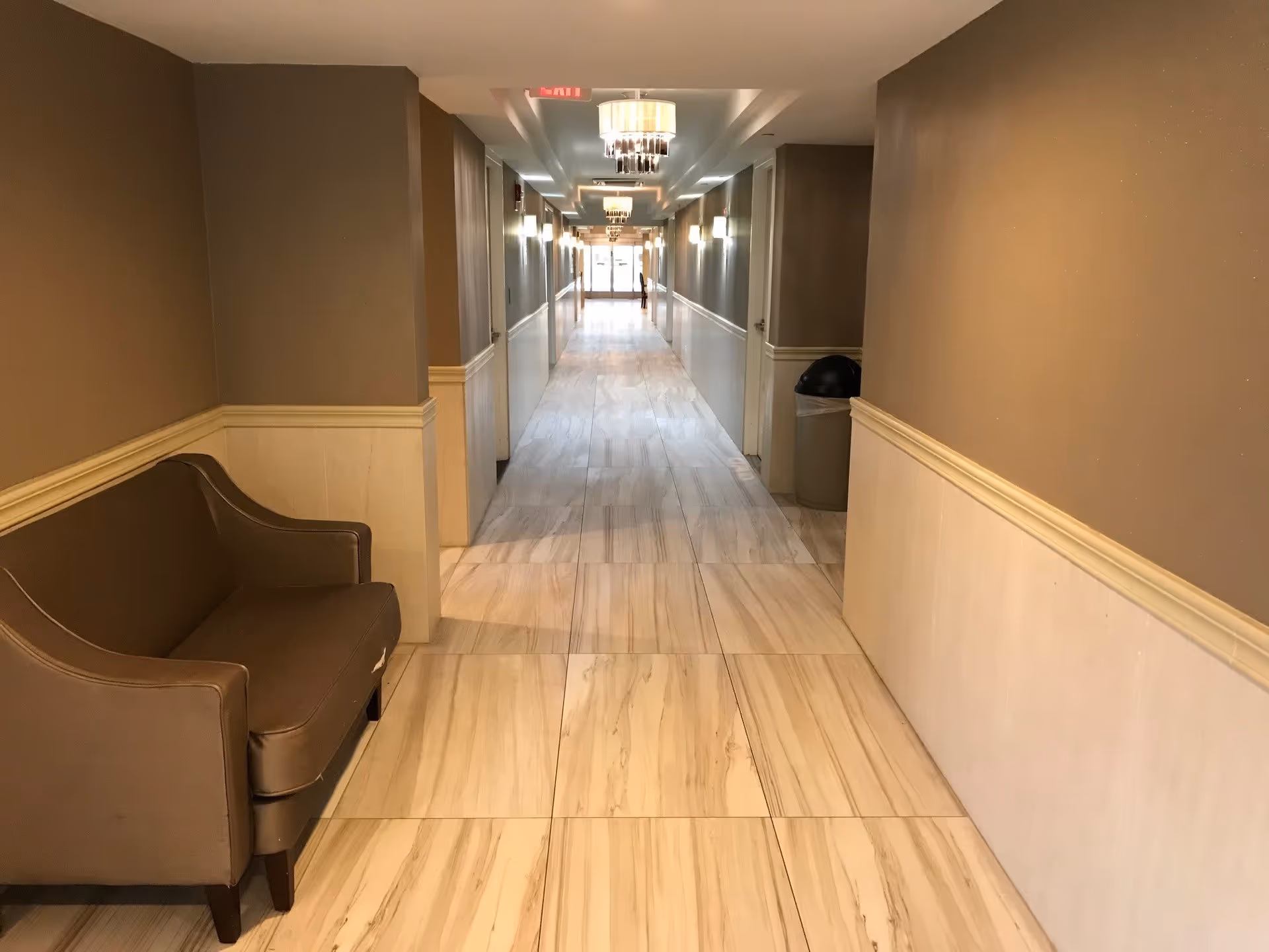 A long, well-lit hallway with beige walls and light-colored tiled floor. There is a brown upholstered bench on the left side and a trash can on the right side. Several ceiling lights and wall sconces illuminate the corridor, which leads to a glass door at the far end.