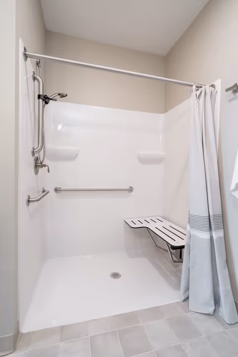 Accessible walk-in shower with grab bars, a fold-down bench, handheld showerhead, and a shower curtain.