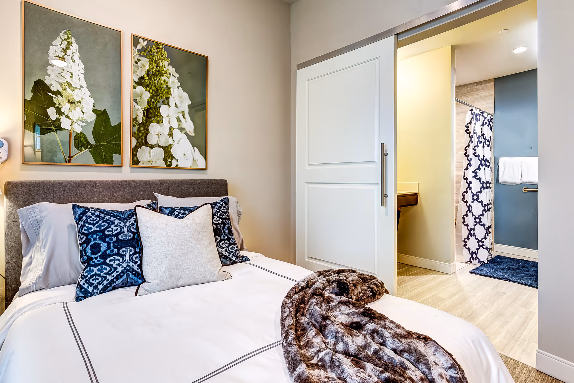 A cozy bedroom with a neatly made bed featuring white and blue patterned pillows and a brown fur throw blanket. Above the bed are two framed floral artworks. To the right, a sliding door opens to a bathroom with a shower curtain, towel rack, and a blue bath mat on the floor.