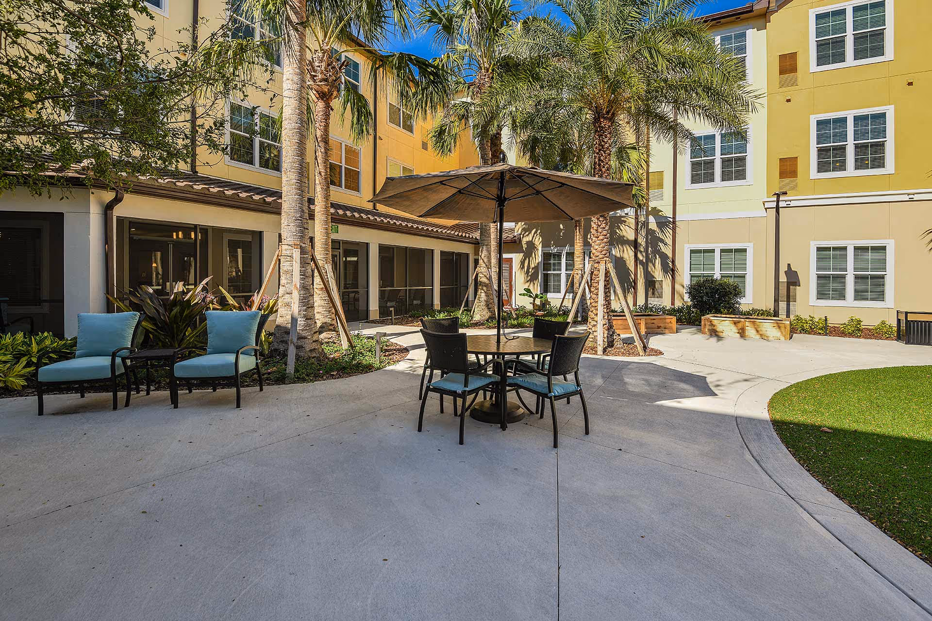 Outdoor courtyard area at Providence Living at Hunter's Creek with patio furniture including a round table with an umbrella and four chairs, two cushioned lounge chairs, palm trees, and a multi-story building with large windows in the background under a clear blue sky.