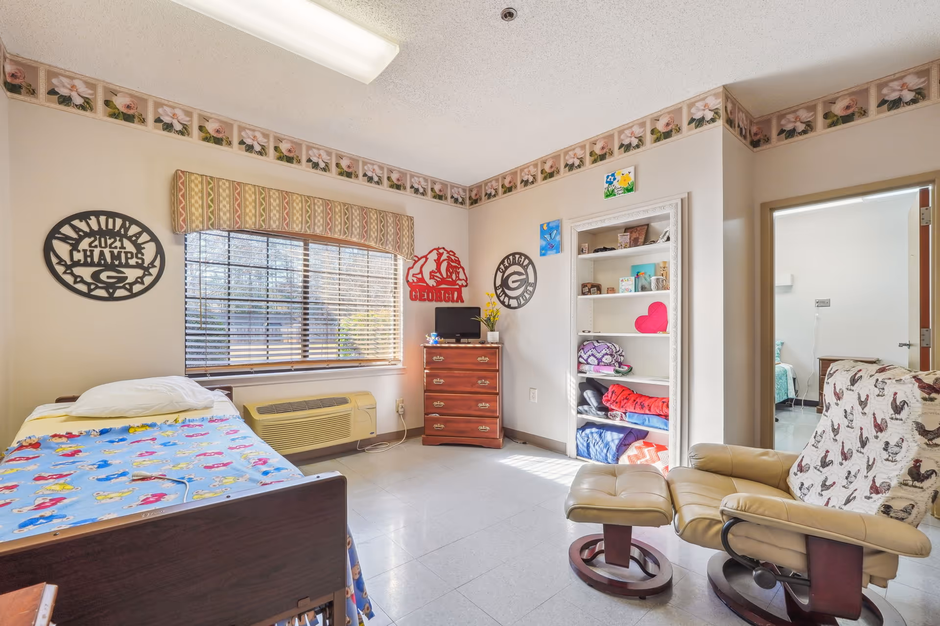 A bright senior living bedroom with a single bed covered in a colorful blanket, a window with blinds and a patterned valance, a wooden dresser with a small TV on top, an open shelving unit with folded blankets and decorative items, and a beige recliner chair with an ottoman covered by a rooster-patterned throw. The walls have floral wallpaper border near the ceiling and sports-themed decorations.