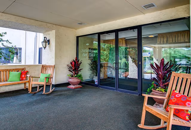 Covered entrance area of Overland Park Place with glass double doors, two large potted plants on either side of the doors, wooden benches and rocking chairs with colorful cushions, and a wall-mounted lantern light.