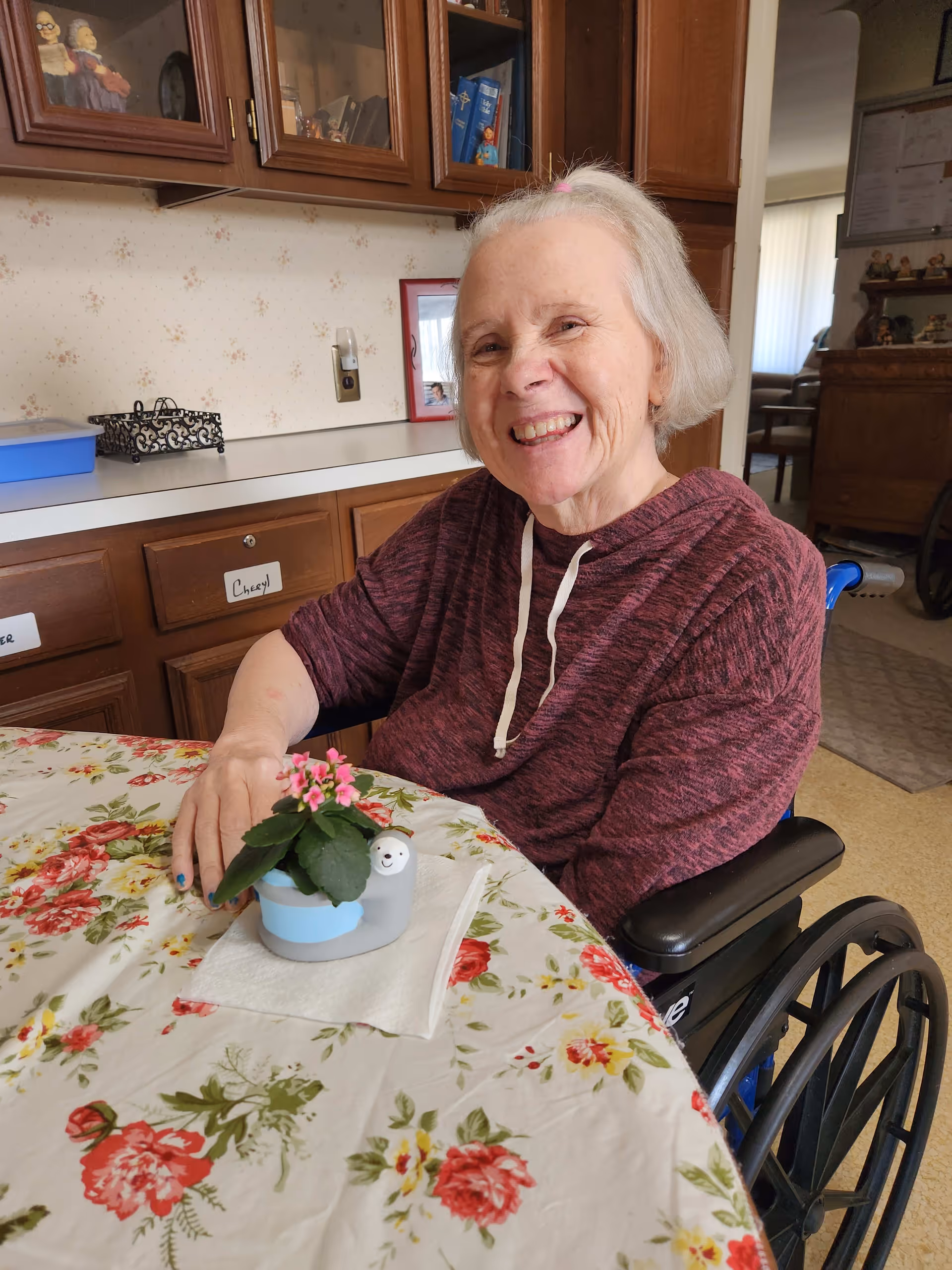 An elderly woman with short gray hair, wearing a maroon hoodie, sitting in a wheelchair at a table covered with a floral tablecloth. She is smiling and resting her hand near a small potted plant with pink flowers in a decorative pot shaped like a smiling snail. Behind her are wooden cabinets and a counter with various items.