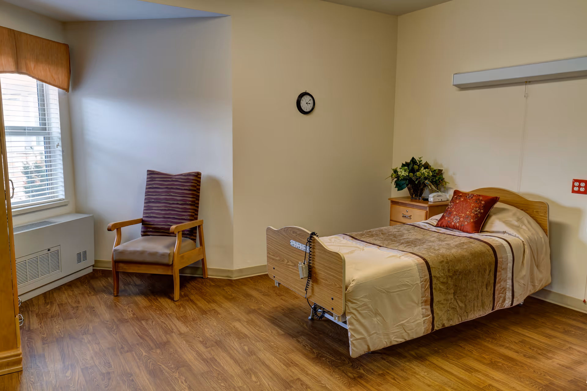 A simple, clean bedroom in a healthcare facility with a single bed made with beige and brown bedding, a red decorative pillow, a wooden nightstand with a telephone and a plant, a wooden chair with a purple cushion near a window with blinds, and a small wall clock above the bed.