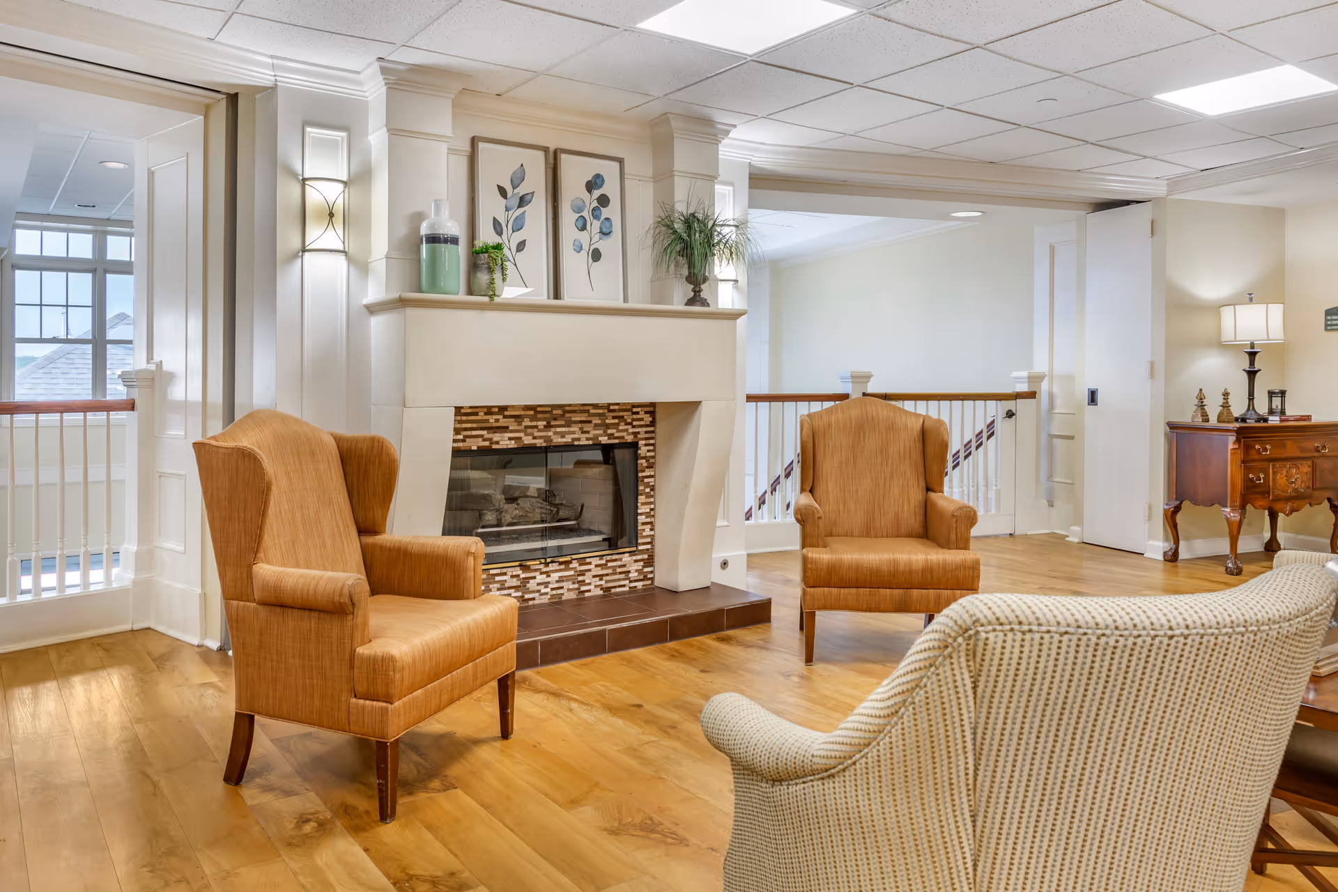 A cozy sitting area in a senior living facility featuring a fireplace with decorative tiles and mantle adorned with vases and framed botanical prints. The room has two orange upholstered armchairs and a beige patterned armchair arranged around the fireplace. Hardwood floors and soft lighting create a warm atmosphere. In the background, there is a wooden side table with a lamp and decorative items, and a railing overlooking a stairway.