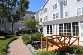 Outdoor courtyard area of a senior living facility with a paved walkway, a small wooden bridge over a dry creek bed, green shrubs, trees, and a multi-story building with many windows in the background under a clear blue sky.